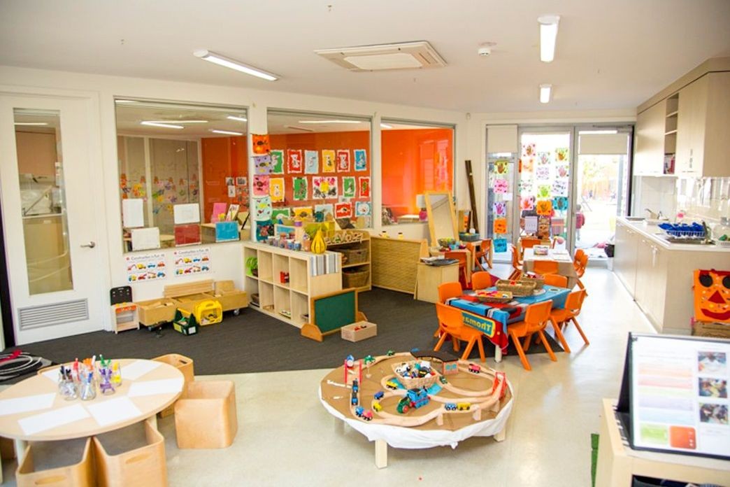 Colorful kindergarten classroom with activity tables, toys, and children's artwork displayed on walls.